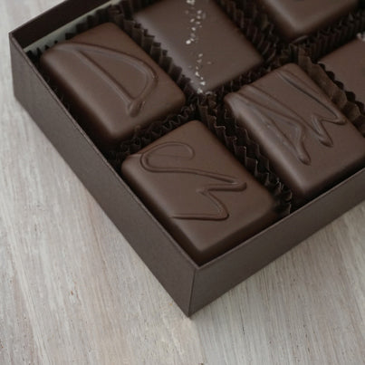 A close up of some of the pieces of chocolate in the dark chocolate assortment. There are four chocolates we can see. The top two are two square pieces. One has a D on it to signify that it is dark chocolate. The other has a line of grey salt going diagonally across. The bottom two are also square. One has ES on it to signify espresso and the other has a DW on it to signify dark with walnuts.