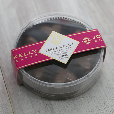 A closed plastic, clear, round, container. The container contains six milk chocolate hearts. The container is held close with a fuchsia label that has a John Kelly Chocolates logo on it.