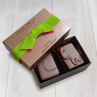 An open two piece assortment. Two chocolates with C and CW labeled on them sit neatly in the box. The light brown lid of the box sits partially on the box. The lid has a lime ribbon across it.