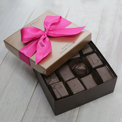 An open 24 piece assortment. Various chocolates are showing from the assortment. The lid of the box is partially on the box and has a fuchsia ribbon on it