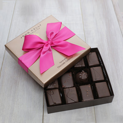 An open 12 piece assortment box. Various chocolates from the assortment as showing. The lid of the box is above the box and has a fuchsia ribbon on it.