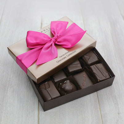 An open eight piece assortment box. A selection of the chocolate from the assortment can be seen. The lid of the box is partially on the box and features a bright fuchsia ribbon.