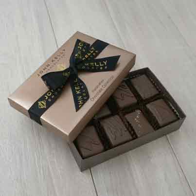An open box of our eight pieces dark assortment. Some of the chocolates can be seen neatly placed in the box. The lid of the box is partially covering some of the chocolates. The light brown lid has a black bow on it that reads John Kelly Chocolates.