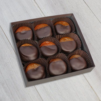 An open box of the chocolate dipped apricots. There are nine total apricots in the box. The apricots are almost completely covered in chocolate leaving a fourth of the top exposed. They are all neatly laid in brown, paper cups.