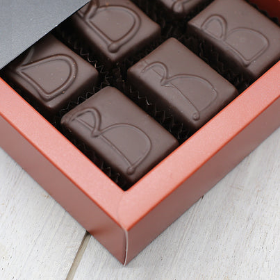 Chocolate with the letter B on them sit in a red box. The chocolates are rectangular in shape.
