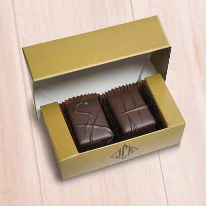 A gold box sits open with two pieces of chocolates in it. One has a CH on it and the other has a H.