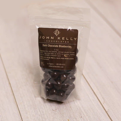 A bag of dark chocolate covered blueberries stands. The bag has a brown label on it with a John Kelly Chocolates logo.