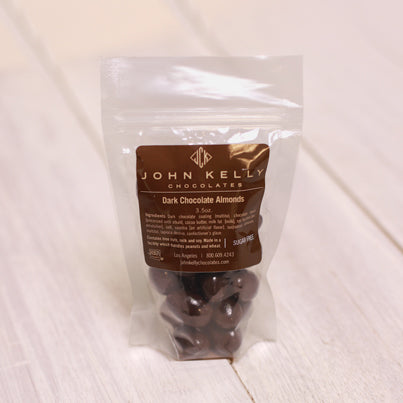 A bag of dark chocolate covered almonds sits. The bag has a brown label on it with a John Kelly Chocolates logo on it.