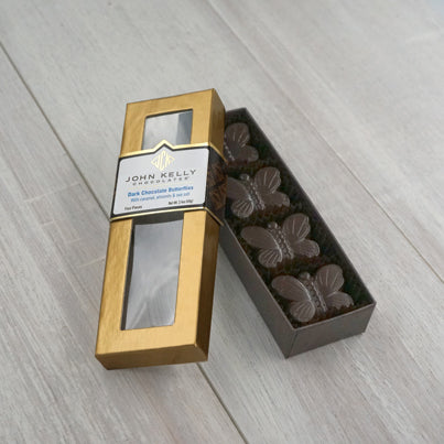 An open fourpiece dark chocolate butterflies box. The box is long and rectangular. The box holds the four butterflies vertically, they are placed neat and snug in the box. The lid of the box is leaning against the box and is gold in color. The lid has a window on it and a white label with a John Kelly Chocolates logo on it.
