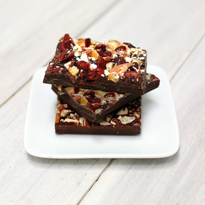 Three bars of the dark chocolate bark stacked vertically. They are topped with bits of orange and cranberry, They are sitting on a white, square plate.