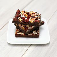 Three bars of the dark chocolate bark stacked vertically. They are topped with bits of orange and cranberry, They are sitting on a white, square plate.