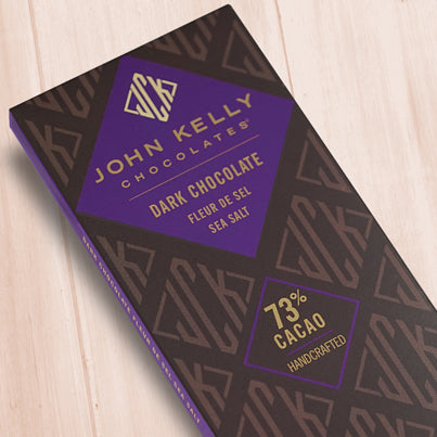 A dark brown, rectangular box. There is a purple diamond on it that holds a John Kelly Chocolates logo and the product name.