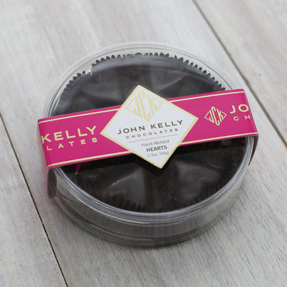 Six dark chocolate hearts are in a closed plastic, clear, round container. The label on the lid is fuchsia and has a John Kelly Chocolates logo on it.