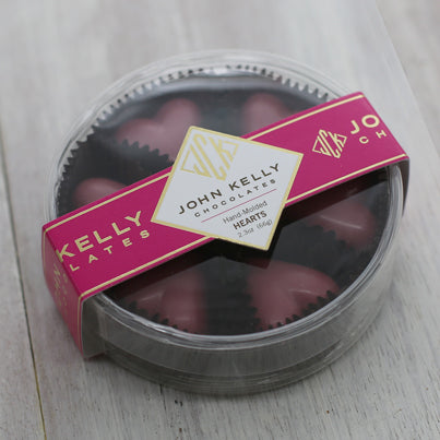 Six pink ruby chocolate hearts sit in a closed container. The container is plastic, clear, and round. A fuchsia label is on the lid and has a John Kelly Chocolates logo on it.