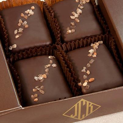Four chocolates sit in a dark, brown box. They all have pinkish salt going diagonally across them.