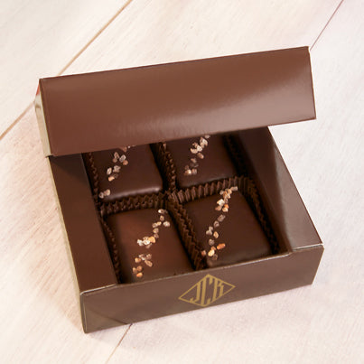 Four chocolates sit in a dark, brown box. They all have pinkish salt going diagonally across them.
