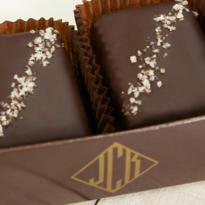 Two grey salted truffle fudge bites in a dark brown box. The box features a gold John Kelly Chocolates logo.