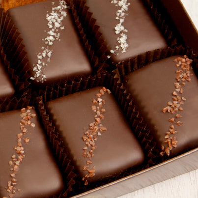 A close up of salted chocolates in a brown box. The salt on top of the chocolates is grey for some and red for others.