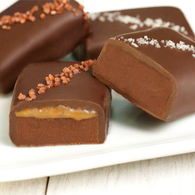 Four salted truffle fudge bites sit on a plate. the two on the left have red salt going diagonally across the chocolate. The other two bars have grey salt on them going diagonally.