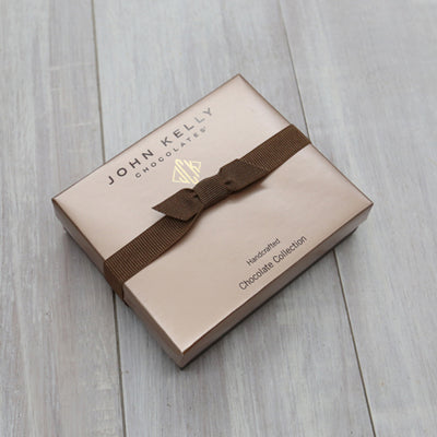 A closed six piece assortment box. The lid of the box is a light brown and has a dark brown ribbon horizontally across it.