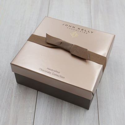 A closed 24 piece assortment box. The box is square and has a dark brown bottom and a light brown lid. A ribbon holds the box closed. The ribbon is a dark brown.