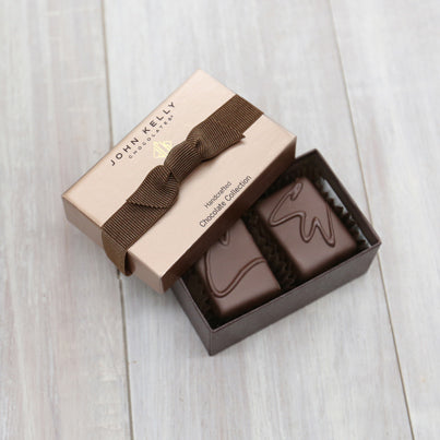 An open two piece assortment. Two chocolates with C and CW labeled on them sit neatly in the box. The light brown lid of the box sits partially on the box. The lid has a brown ribbon across it.