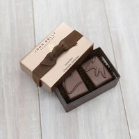 An open two piece assortment. Two chocolates with C and CW labeled on them sit neatly in the box. The light brown lid of the box sits partially on the box. The lid has a brown ribbon across it.