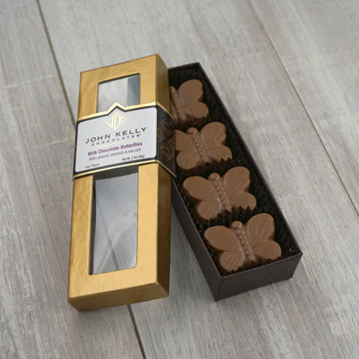 An open box of the four piece milk butterflies. The bottom of the box is brown and holds four milk chocolate butterflies sitting vertically in the long, rectangle box. The lid of the box is gold with a window. The label of the box sits three quarters of the way up the window and has a John Kelly Chocolates label on it.