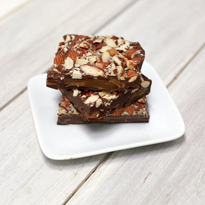 Three bars of the milk chocolate bark sit stacked vertically. They are filled with almonds and caramel. They are sitting on a white, square plate.