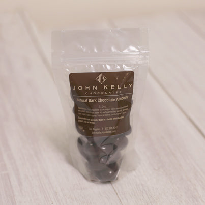 A bag of dark chocolate almonds sits closed. There is a brown label on the bag with a John Kelly Chocolates logo on it.