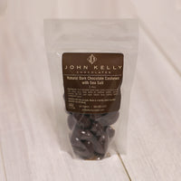A bag of chocolate covered cashews stands. The label on the bag is brown and features a John Kelly Chocolates logo.