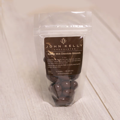 A bag of milk chocolate almonds stands with a brown label on it. The label features a John Kelly Chocolates logo on it.
