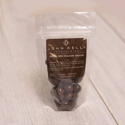 A bag of milk chocolate almonds stands with a brown label on it. The label features a John Kelly Chocolates logo on it.