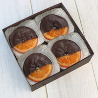 A open with four chocolate covered orange slices in it. The orange slices are sitting in white, paper cups. The orange slices are covered in chocolate, leaving one fourth of the orange slice uncovered.