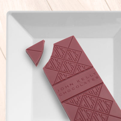 A pink ruby chocolate bar on a white, square plate. The bar is missing the top left corner of the bar. The bar is molded with the John Kelly Chocolates logo all over it.