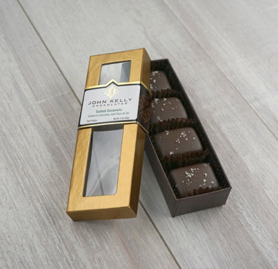 An open four piece fleur de sel salted caramels box. The box is long and rectangular, it holds the four caramels vertically. The lid of the box is leaned against the box and is gold with a window. The label on the lid is white and has a John Kelly Chocolates logo on it.