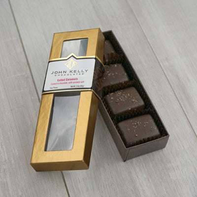 An open box of the four piece applewood smoked salted caramels. The box is long and rectangular and has the caramels sitting vertically in the box. The lid of the box is leaned against the box. The lid is gold and has a window on it. The label is three quarters of the way up the lid and is white with a gold John Kelly Chocolates logo on it.