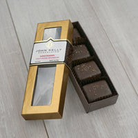 An open box of the four piece applewood smoked salted caramels. The box is long and rectangular and has the caramels sitting vertically in the box. The lid of the box is leaned against the box. The lid is gold and has a window on it. The label is three quarters of the way up the lid and is white with a gold John Kelly Chocolates logo on it.