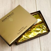 Two gold foiled wrapped rectangle bars sit in a brown box. A gold lid sits partially on the box.