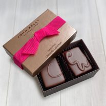 An open two piece assortment. Two chocolates with C and CW labeled on them sit neatly in the box. The light brown lid of the box sits partially on the box. The lid has a fuchsia ribbon across it.