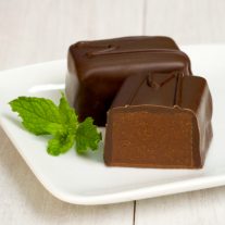 Two mint chocolate pieces sit on a plate. One of them is cut revealing the chocolate-y inside. A sprig of mint sits beside it.