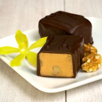 Two pieces of chocolate sit on a plate. One is cut open revealing the white chocolate vanilla inside as well as walnuts. A vanilla flower sits on the left and a walnut sits to the right of the chocolate