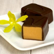 Two pieces of chocolate sit on a plate. One is cut open revealing the white chocolate vanilla inside. A vanilla flower sits on the left and a walnut sits to the right of the chocolate