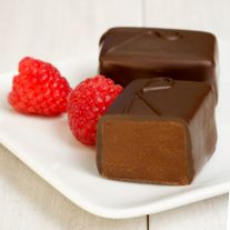 Two raspberry chocolate pieces sit on a white plate. One is cut open revealing the chocolate inside. Two raspberries sit to the left of the chocolate.