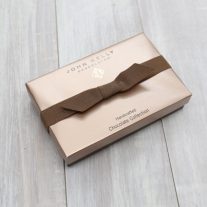 A closed eight piece assortment box. The box is similar in shape to a rectangle and has a light brown lid on it. A brown ribbon sits horizontally on the box,.