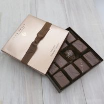 A partially opened box of the 12 Piece Signature Handcrafted Chocolate Collection. Some chocolates from the assortment can be seen. A light brown lid sits partially on the box and had a brown ribbon sitting horizontally around it.