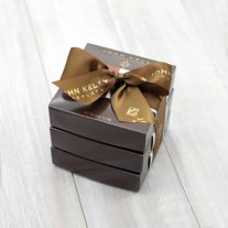 Three dark brown boxes of the same size are stacked vertically. They are tied together with a brown ribbon that reads John Kelly Chocolates.