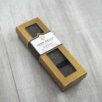 A closed four piece applewood smoked salted caramel box. The box is long and rectangular, with a gold lid. The lid has a window on it where the caramels can be seen. The label on the box sits three quarters of the way up the box and is white. The label includes a gold John Kelly Chocolates logo on it.