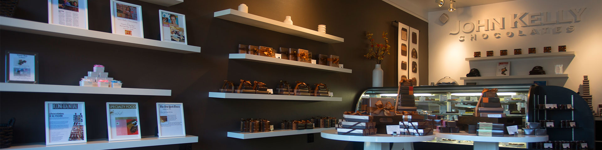 A picture of the Santa Monica store. A brown wall houses shelves of chocolate, while two round tables also hold chocolate.