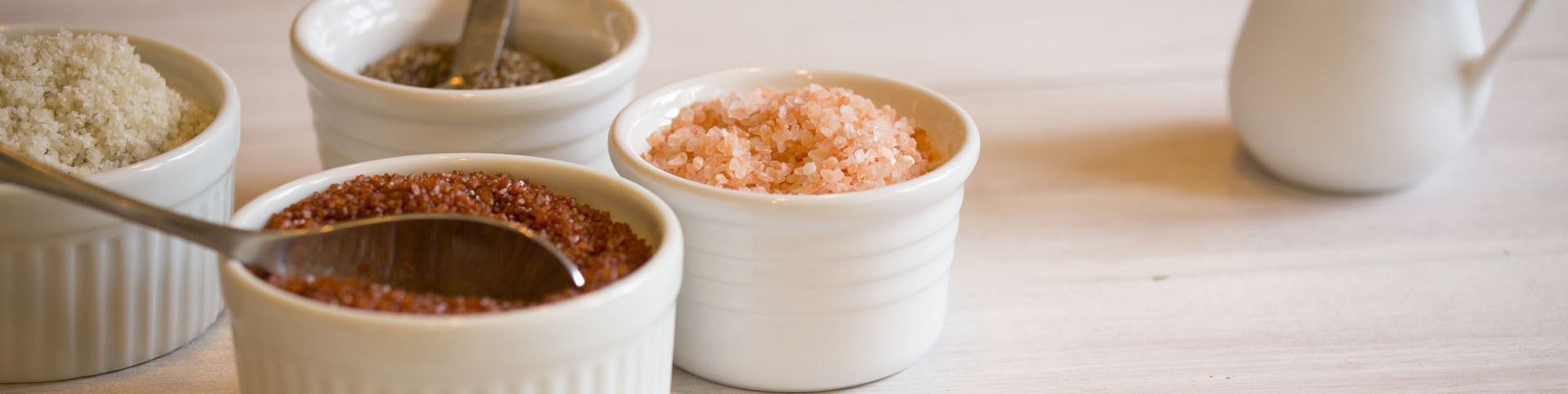 Different salts and spices in white bowls. One of the front bowls has a spoon in it.